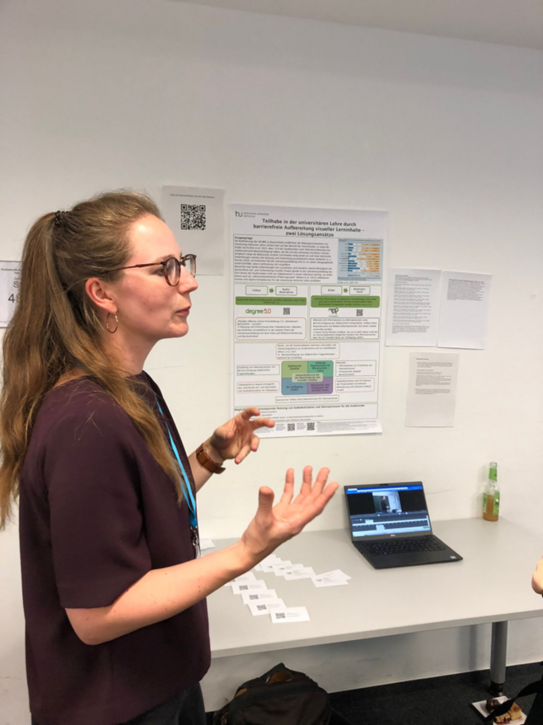 Foto. Leevke Wilkens steht gestikulierend vor einem Poster. Unter dem Poster steht ein Tisch mit einem aufgeklappten Laptop.